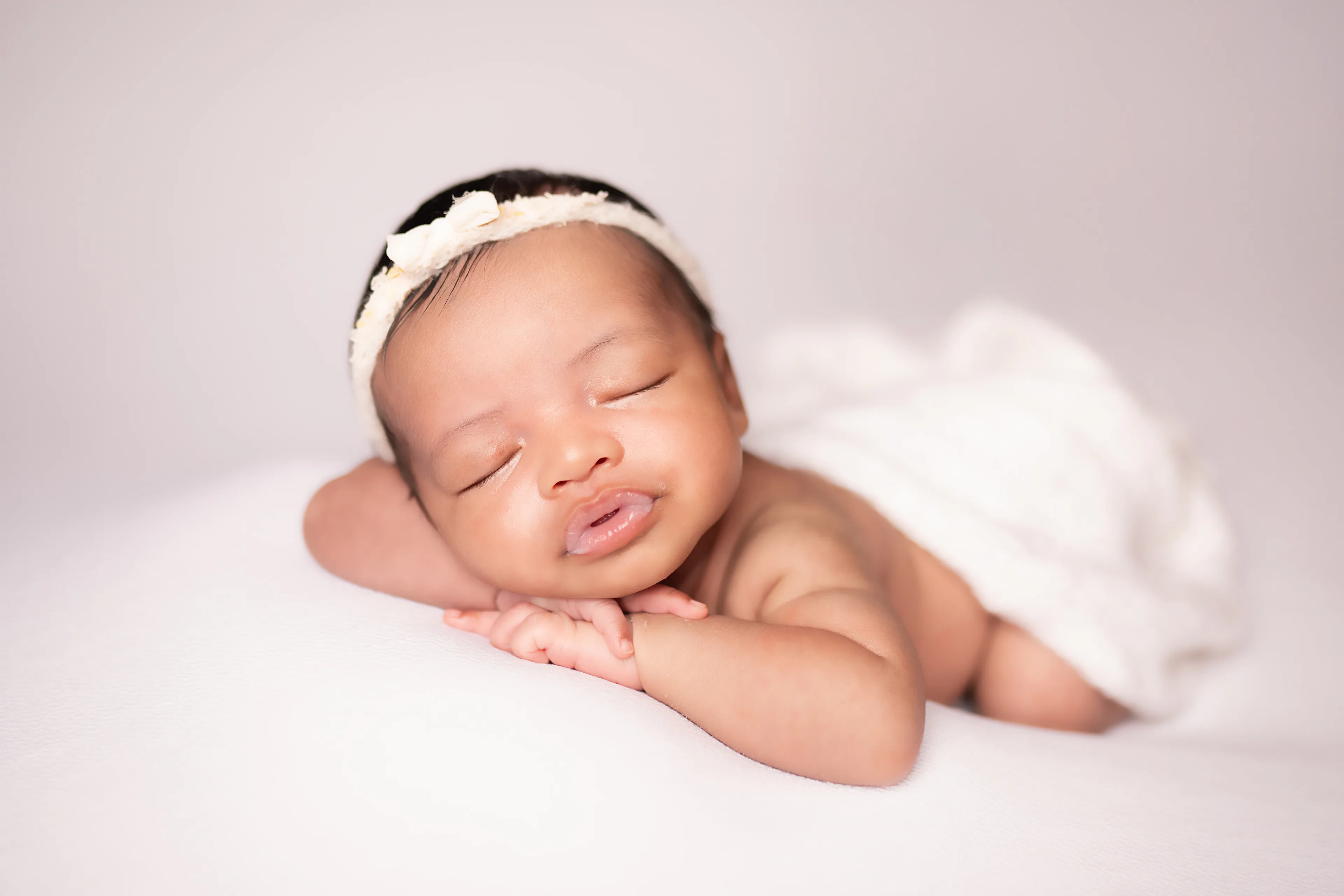 Newborn sleeping on her side on a cream blanket, wearing a knit bonnet, a white wrap draped over her back