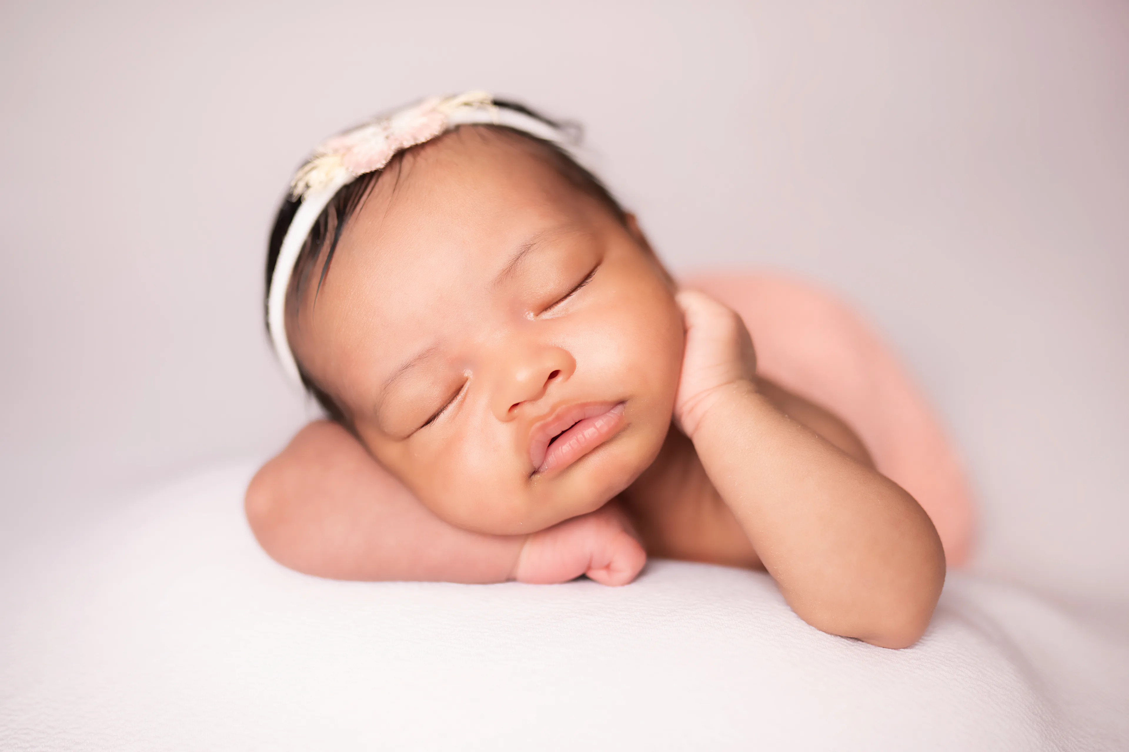 Newborn asleep with her cheek resting on her hand, wrapped in pink on a cream blanket
