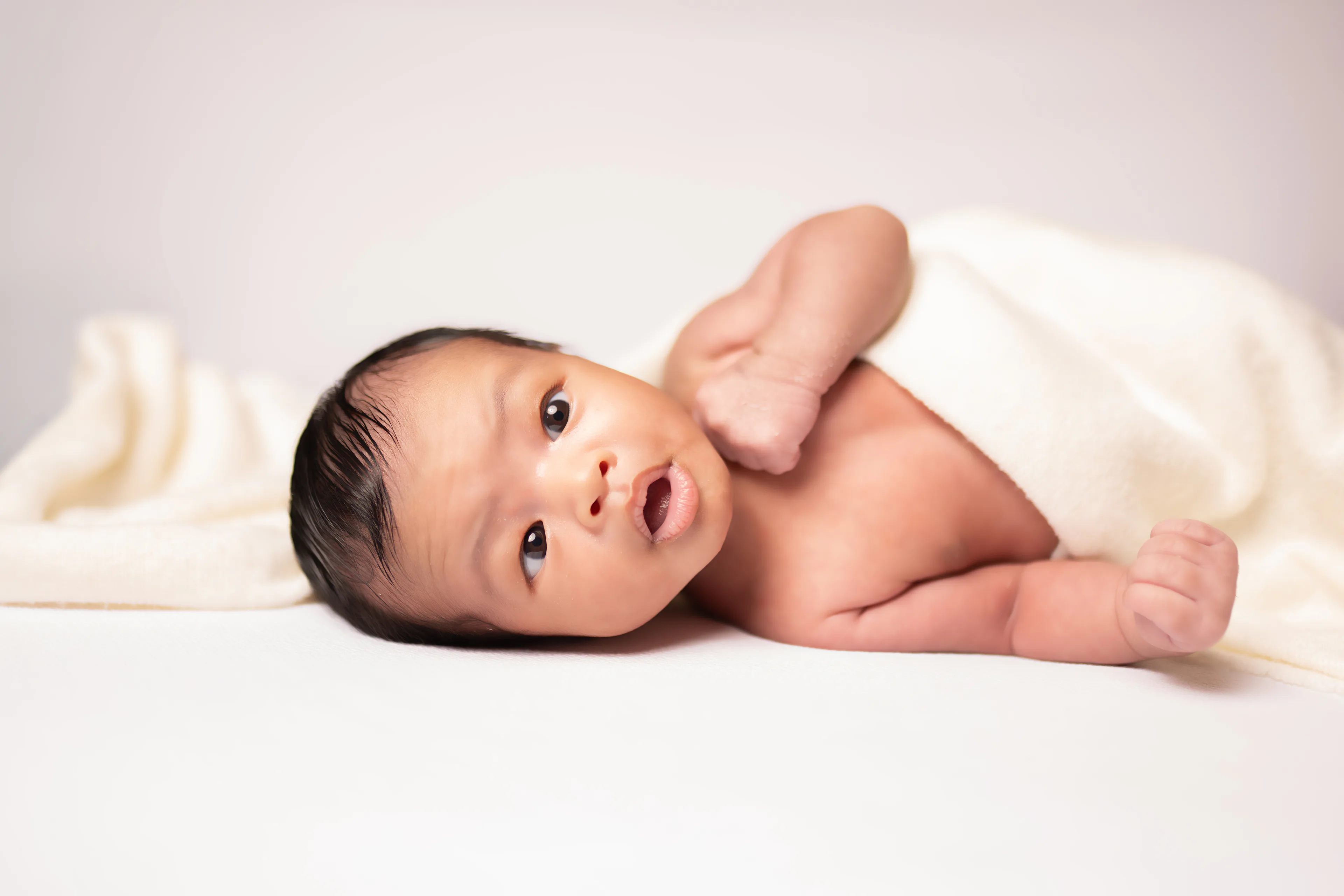 Newborn awake on a white blanket with a cream wrap, one hand raised beside her head
