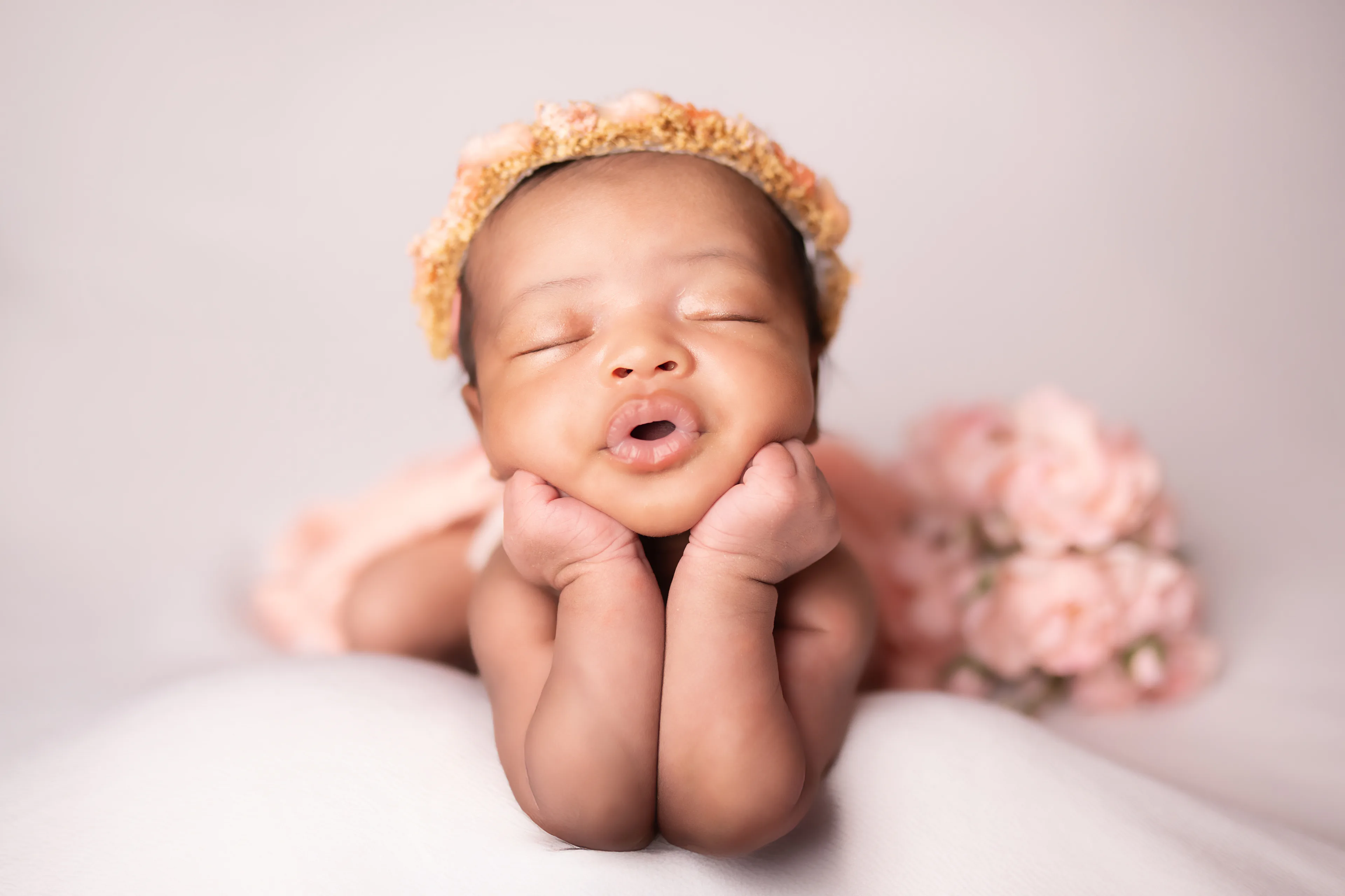 Newborn asleep with her chin propped on both hands, wearing a yellow floral crown, pink blossoms beside her