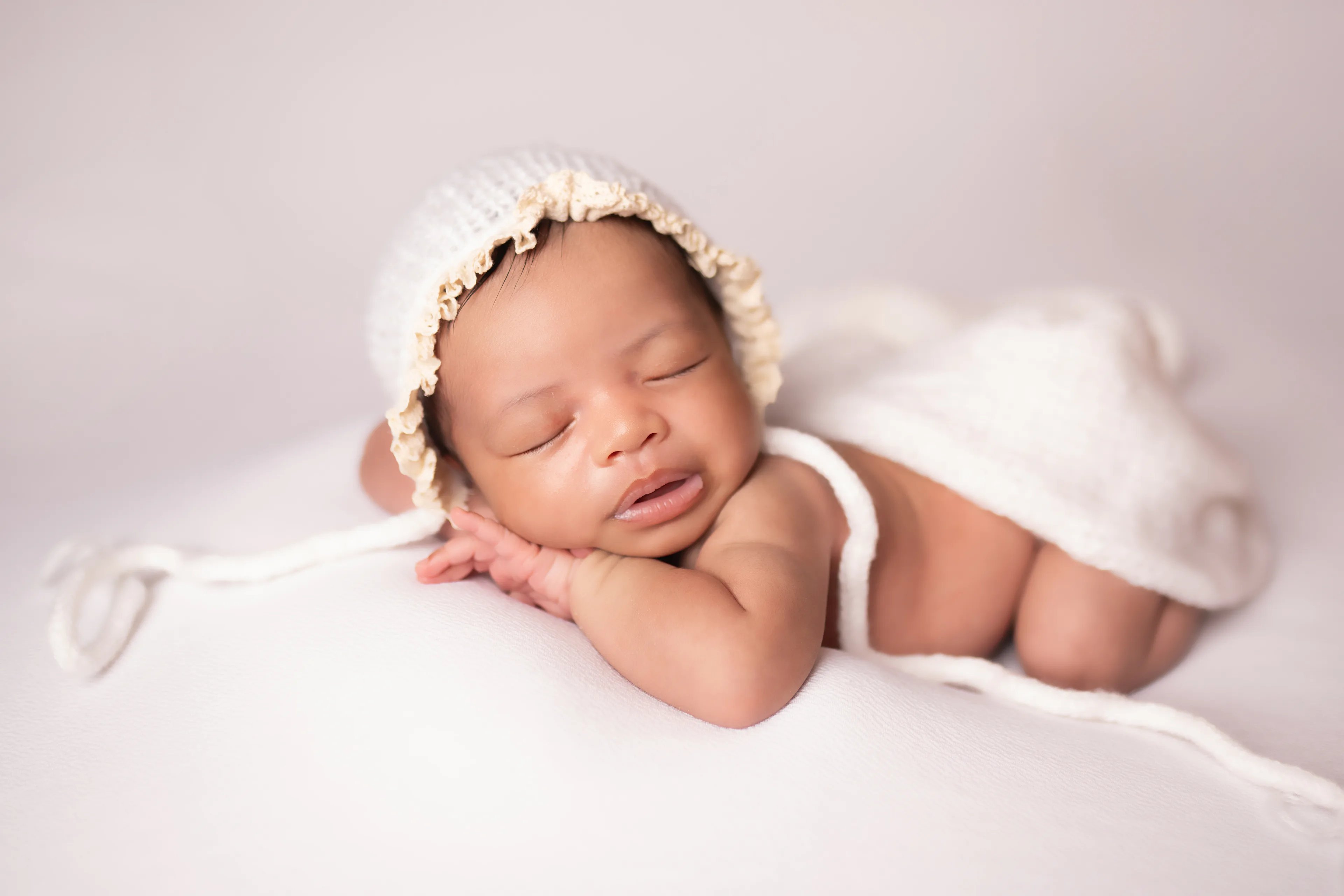 Newborn asleep on a blush blanket in a knit bonnet, wrapped in white, seen from the side