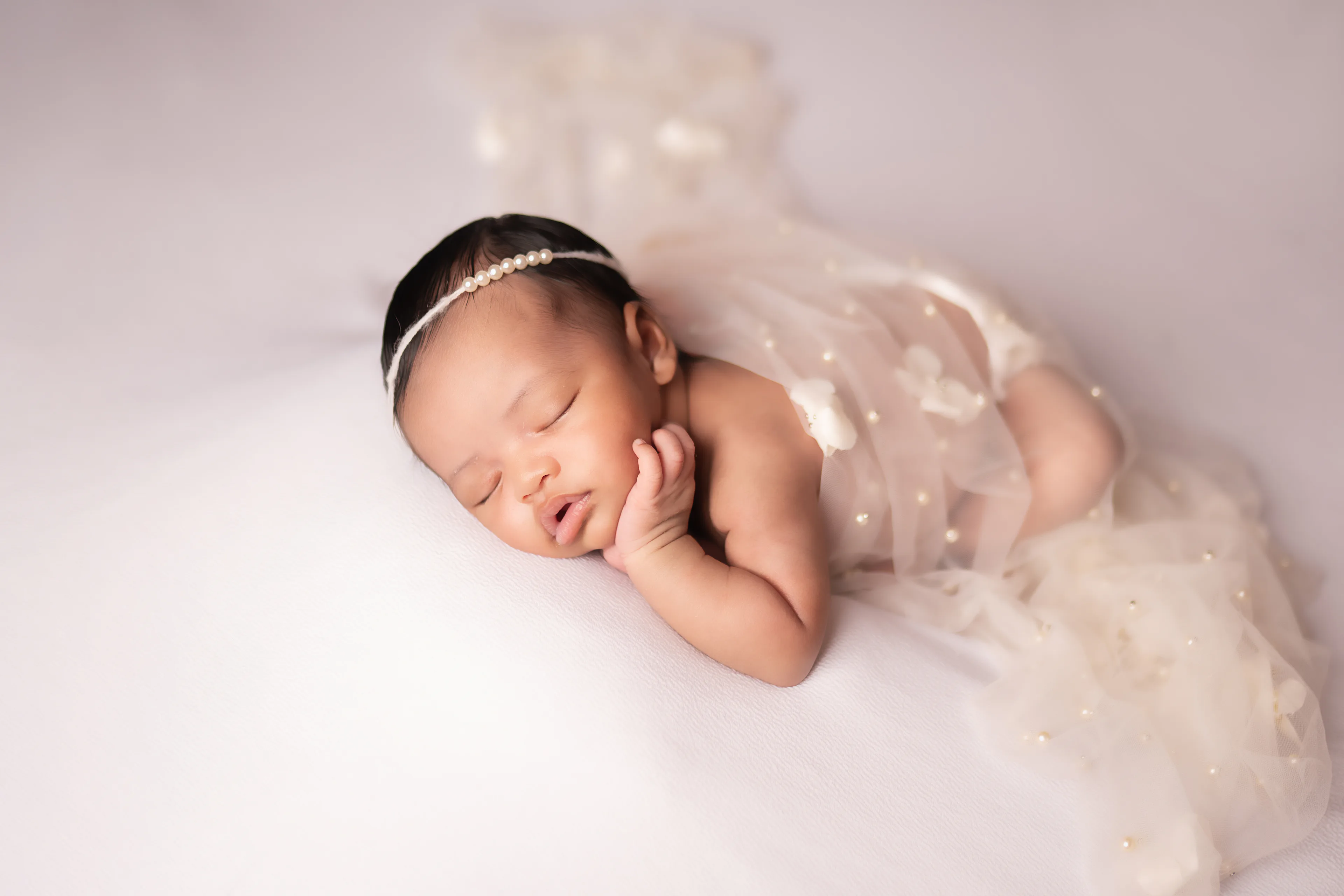 Newborn asleep beneath a sheer pearl-studded veil on a white blanket, her hand resting near her cheek