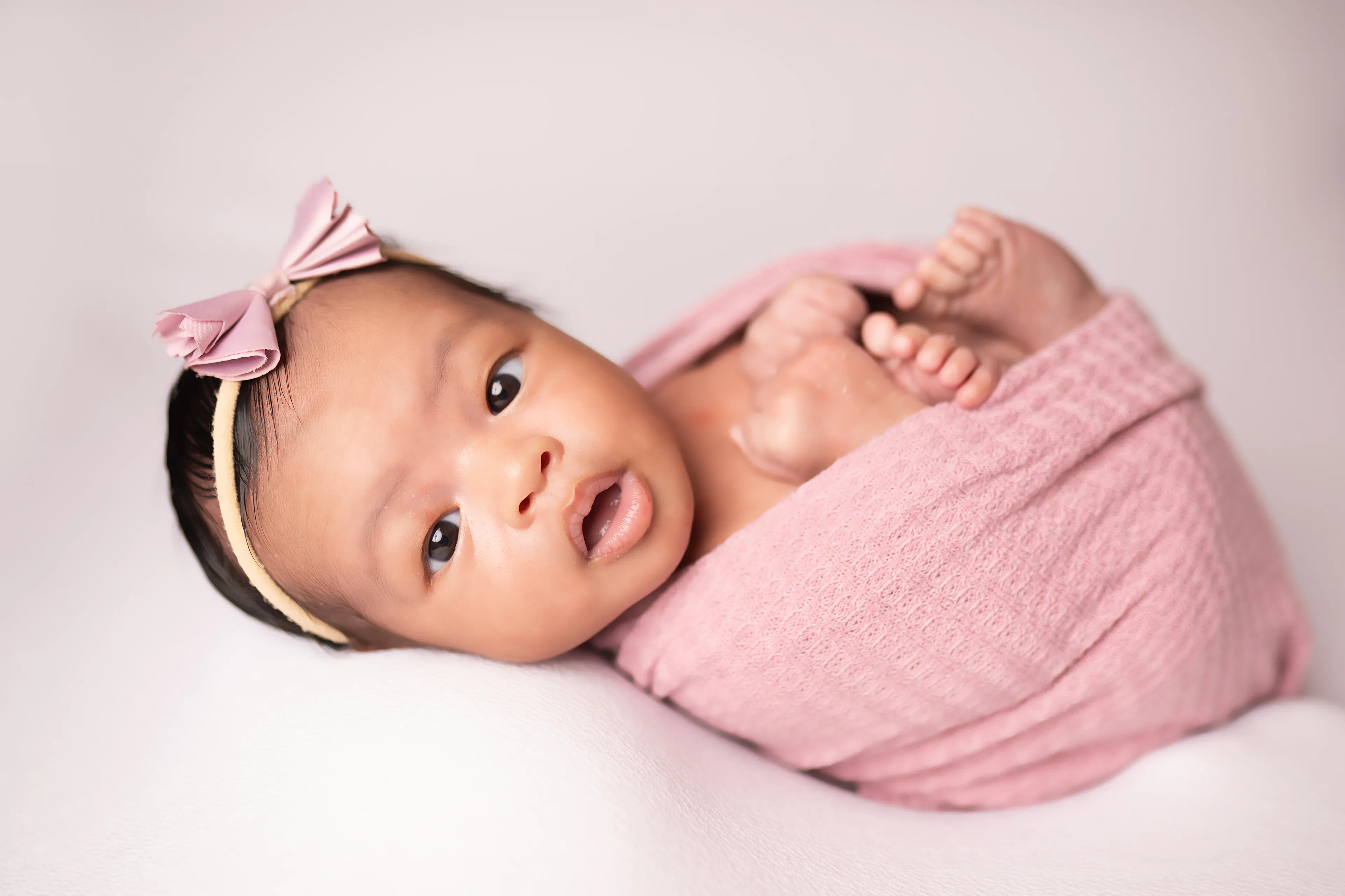 Newborn awake and looking toward the camera, wrapped in a pink knit with a pink bow headband