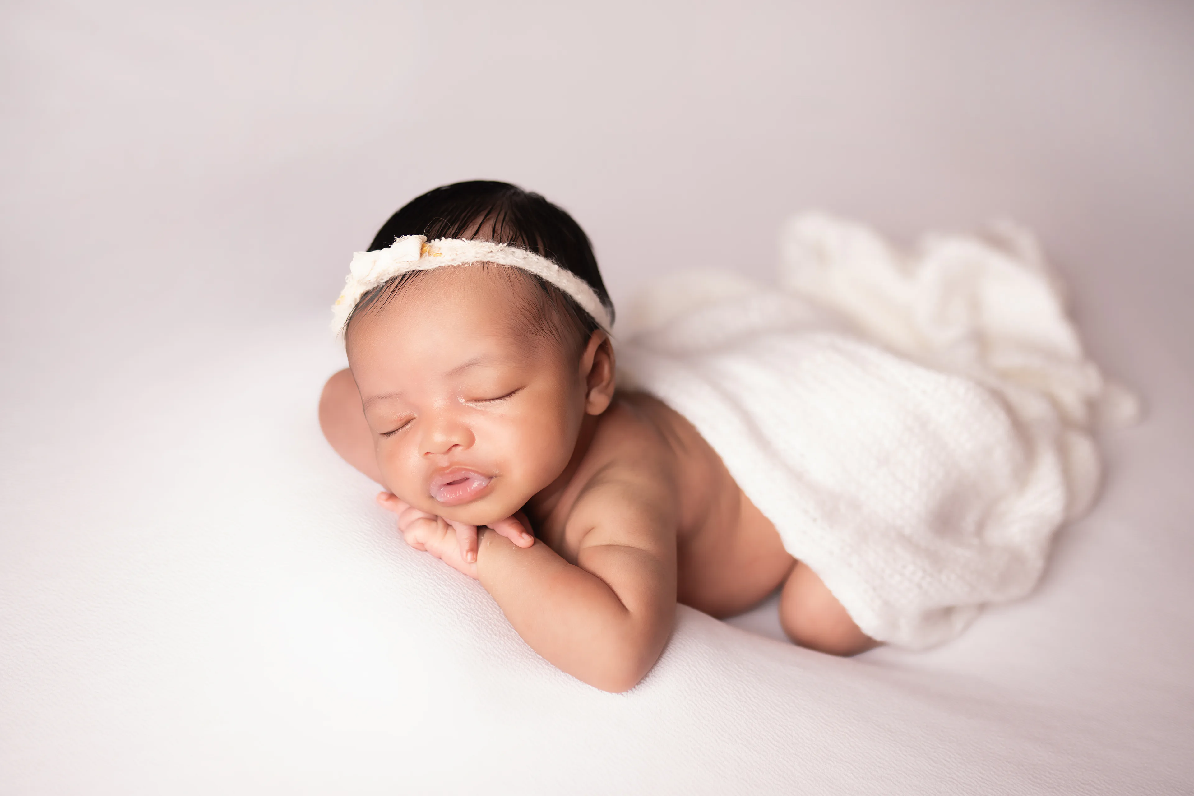 Newborn asleep on a cream blanket in a knit bonnet, hands tucked under her cheek, wrapped in white