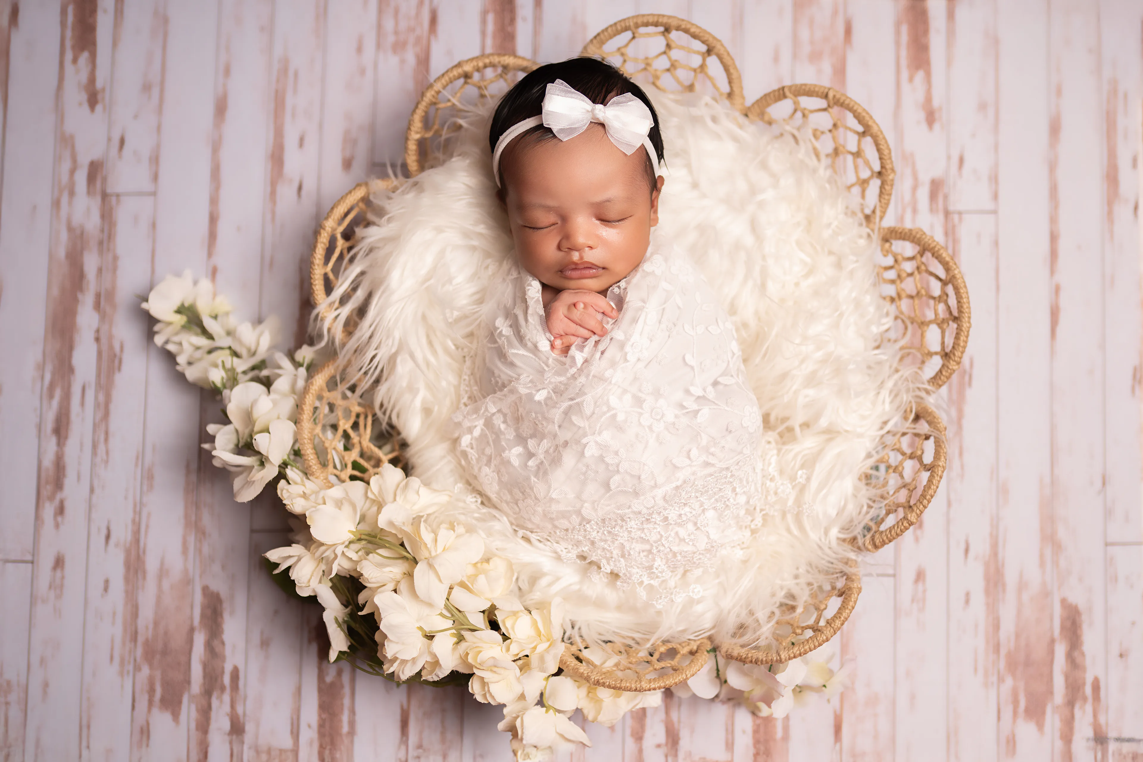 Newborn asleep in a wicker basket lined with cream fur, surrounded by white flowers, on whitewashed wood