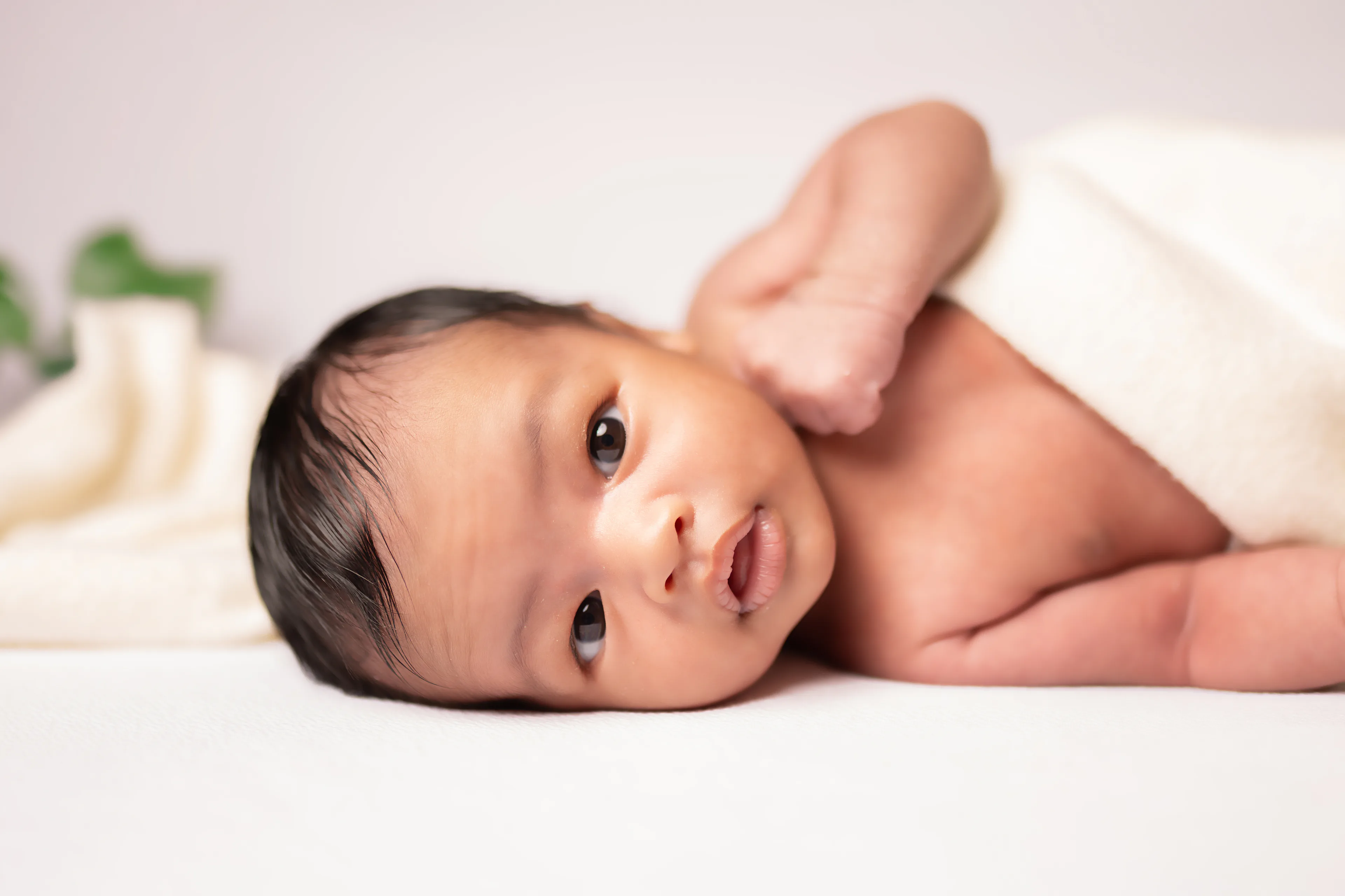 Newborn awake and looking at the camera, lying on a white blanket with a cream wrap