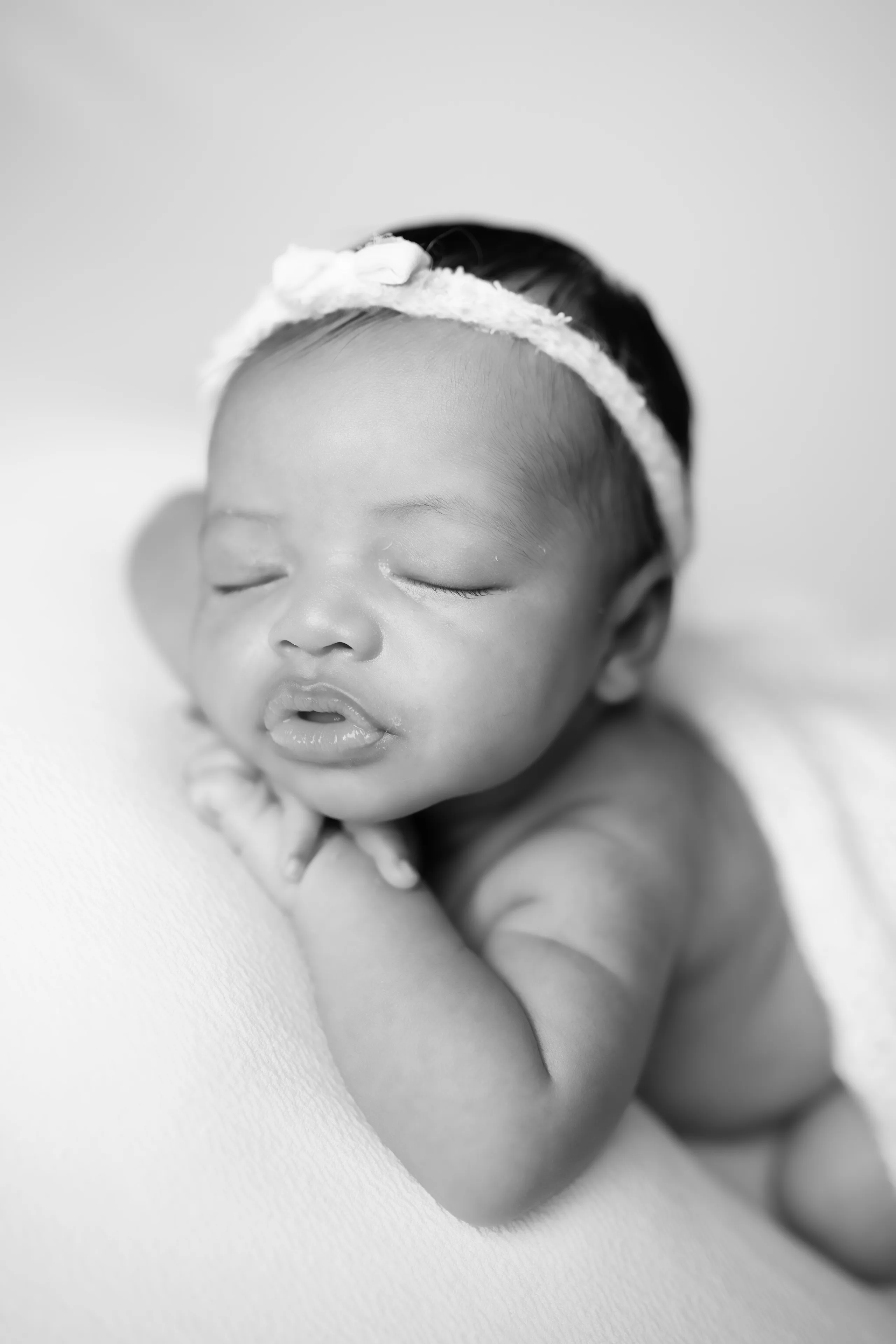 Black-and-white portrait of a newborn asleep in a bonnet, chin resting on her hand