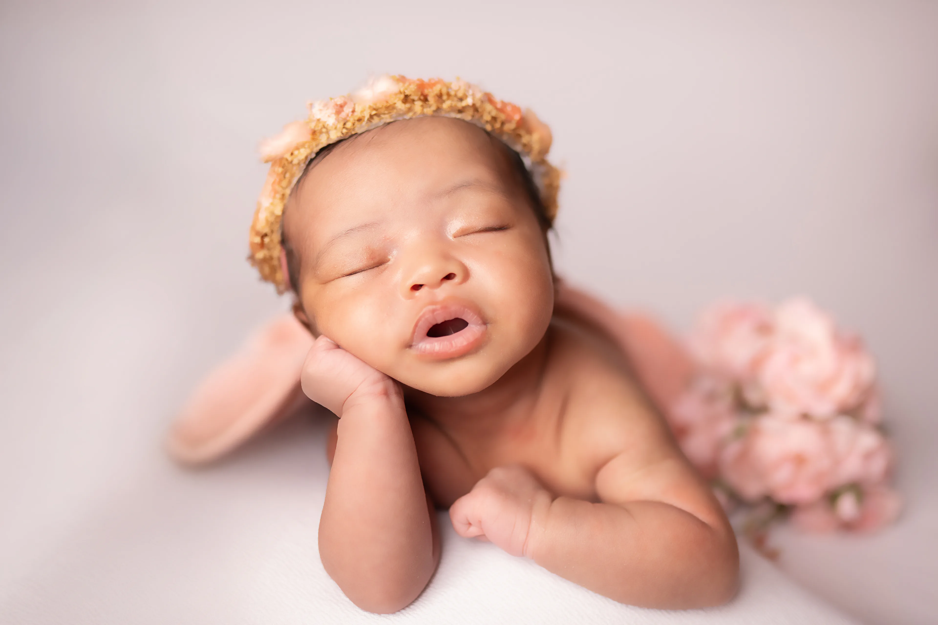 Newborn yawning with her chin on her hands, wearing a yellow floral crown, pink blossoms beside her