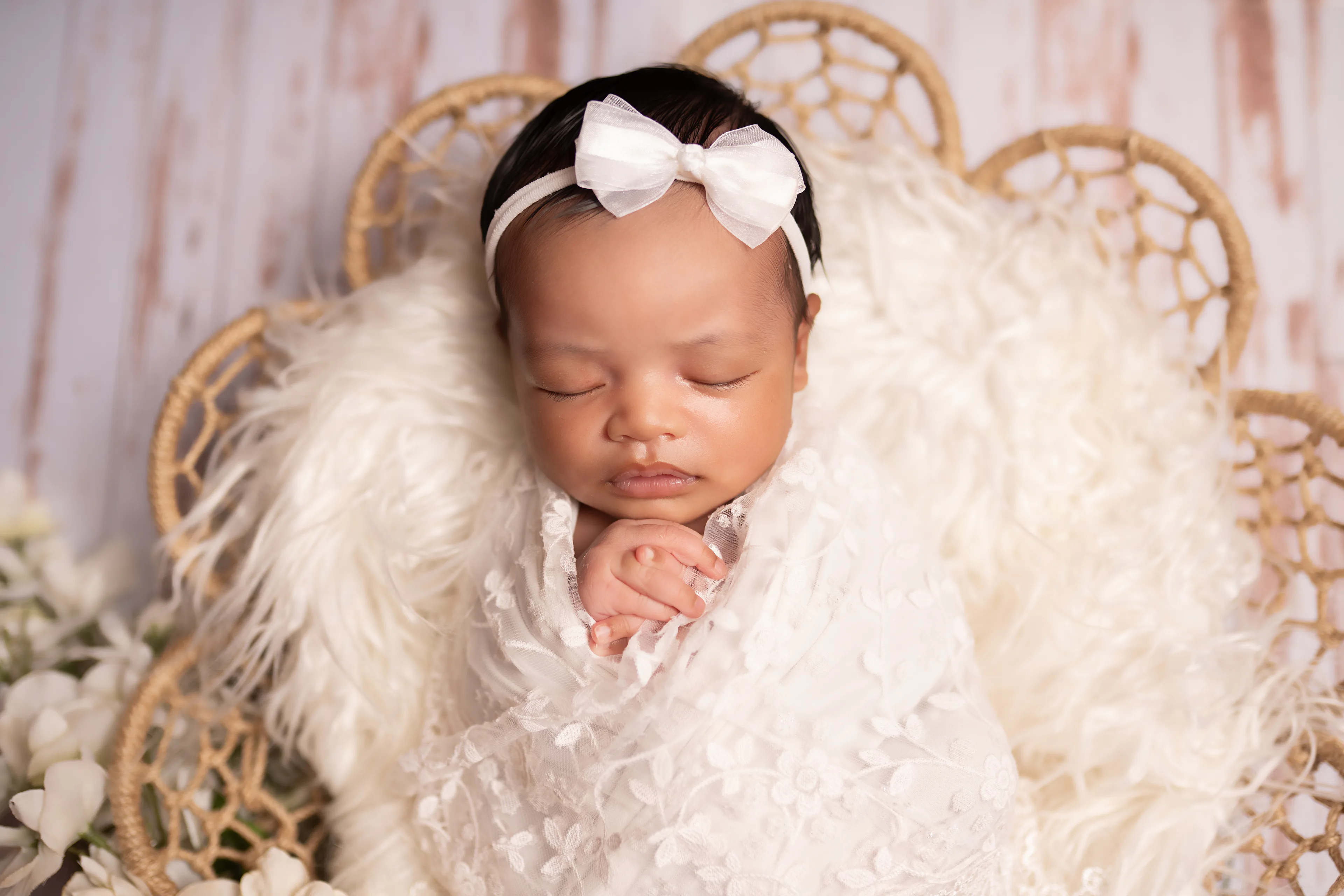 Newborn asleep in a wicker basket lined with cream fur, wearing a white bow, on whitewashed wood
