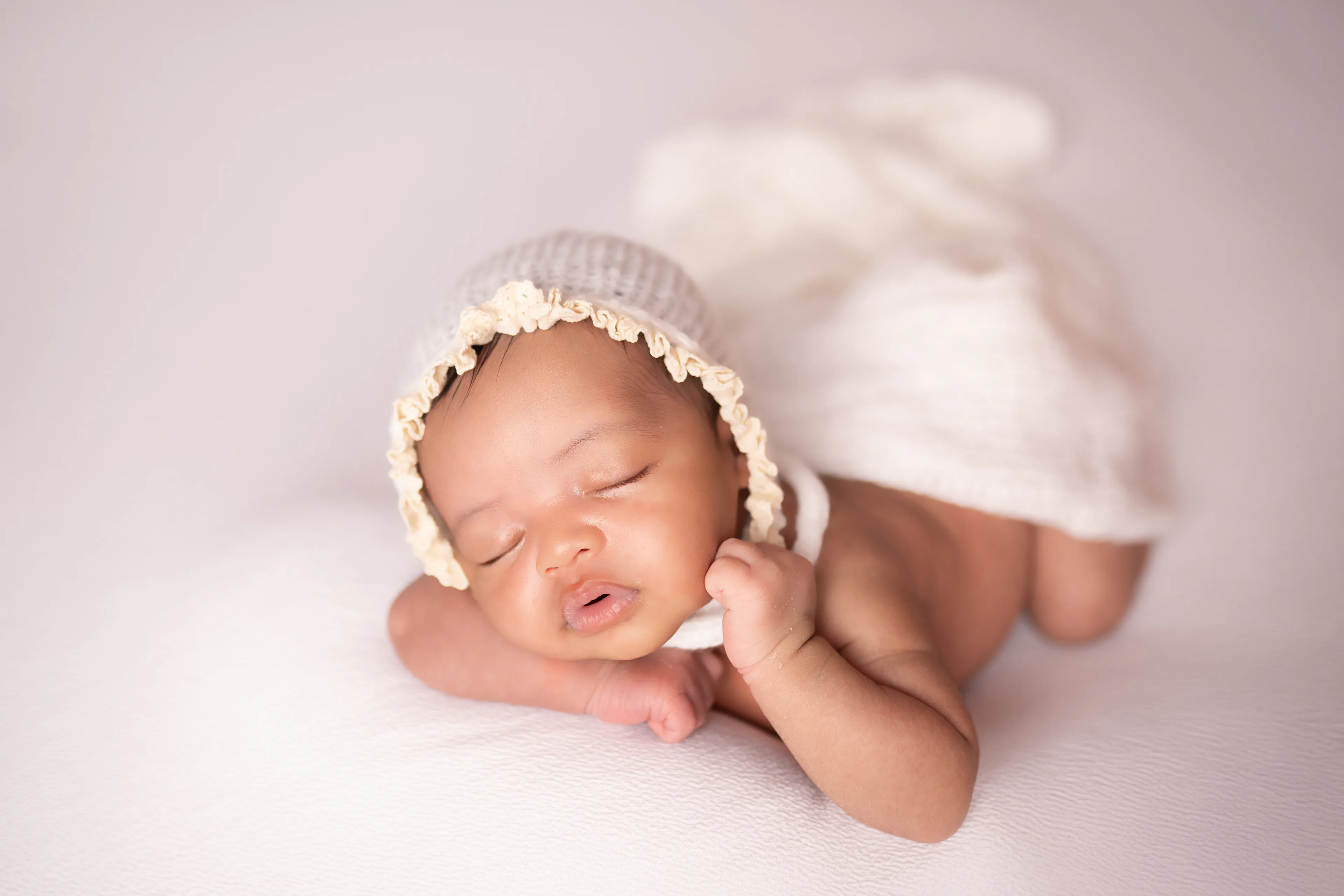 Newborn asleep in a knit bonnet with her hand resting near her chin, wrapped in white