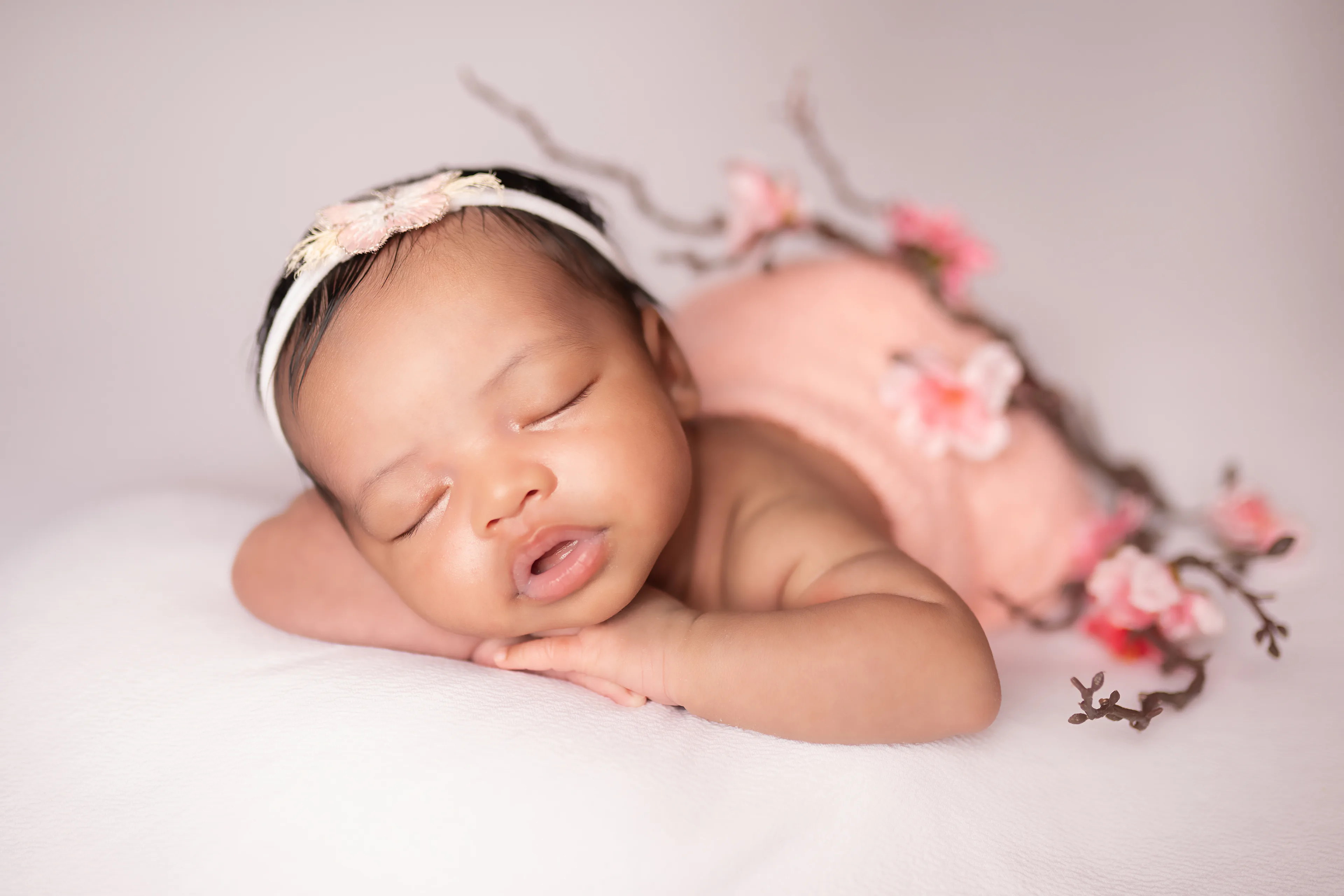 Newborn asleep on a cream blanket with a pink wrap and a branch of cherry blossoms beside her