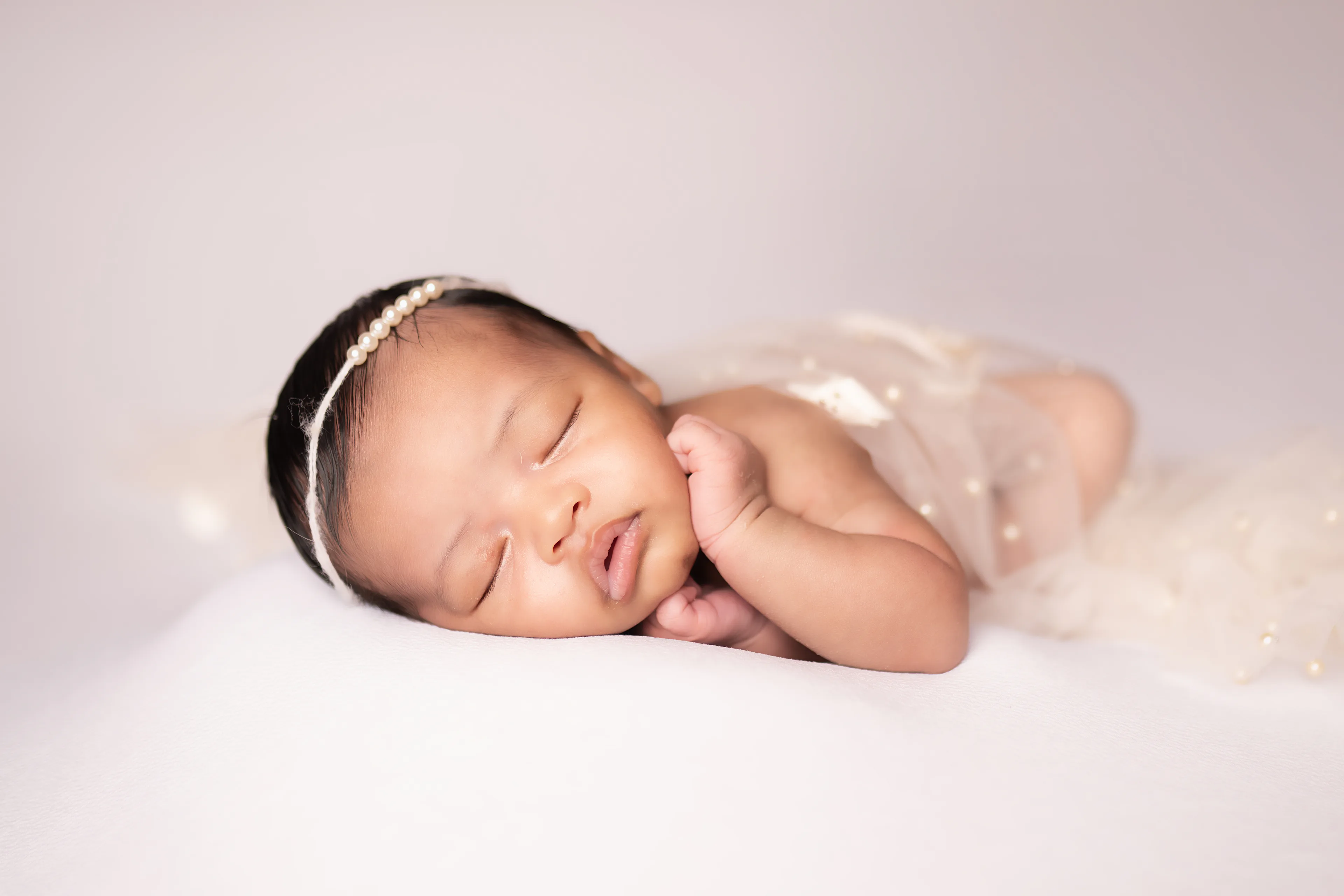 Newborn asleep on a white blanket beneath a sheer pearl-studded veil, wearing a pearl headband