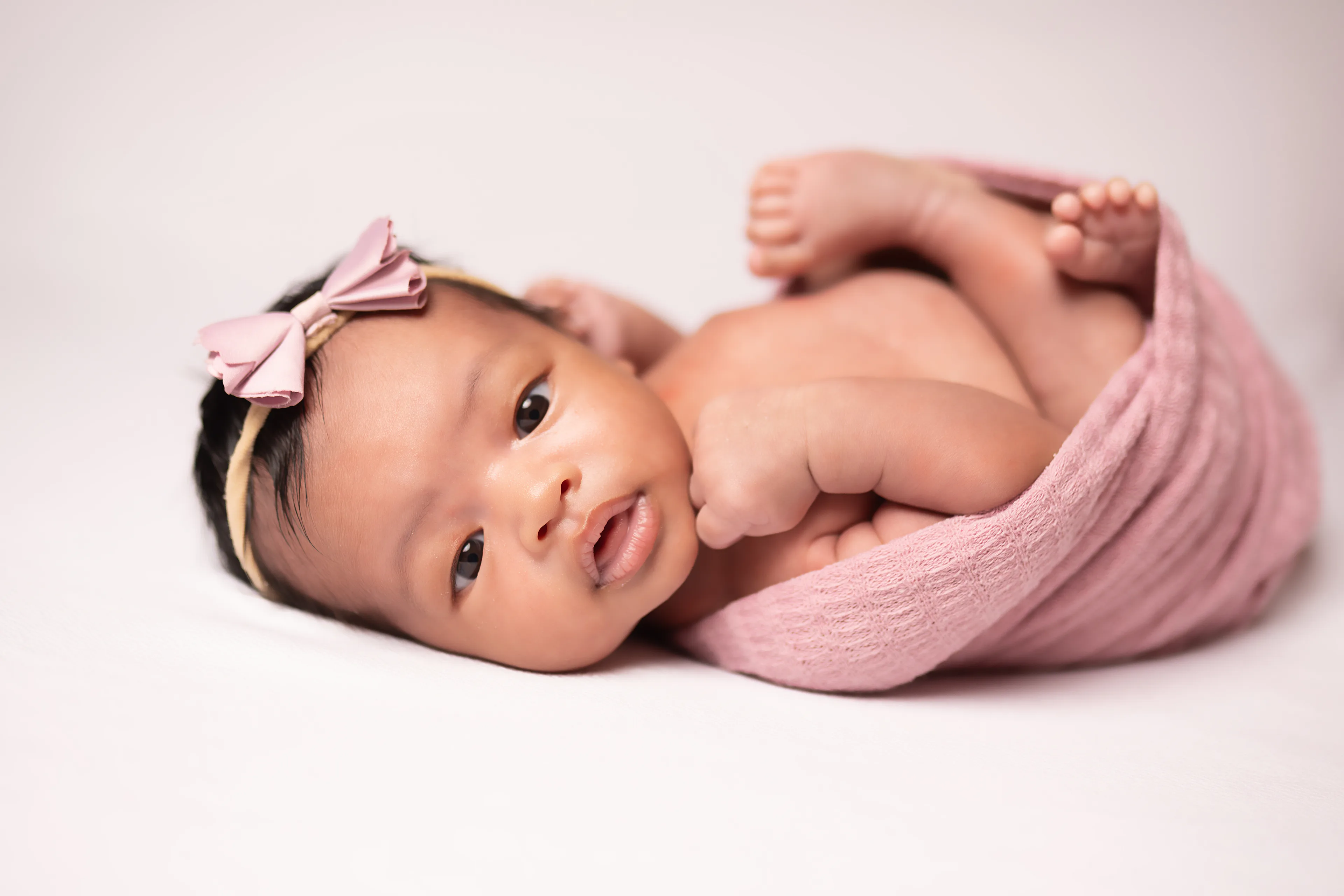 Newborn lying on a white blanket, wrapped in pink with her legs peeking out, eyes open