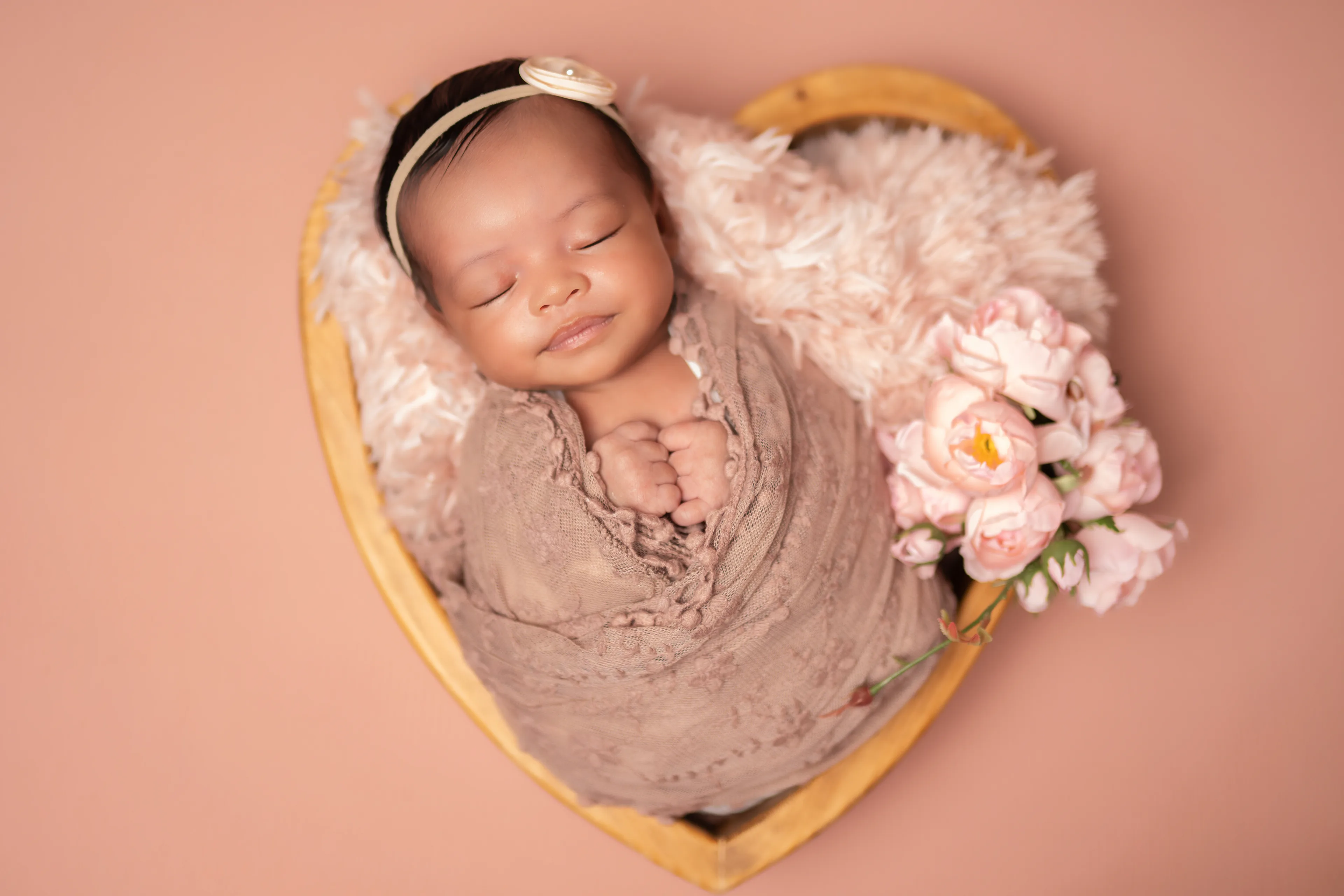Newborn asleep in a heart-shaped wooden bowl with pink blossoms, wrapped in brown, on a peach backdrop