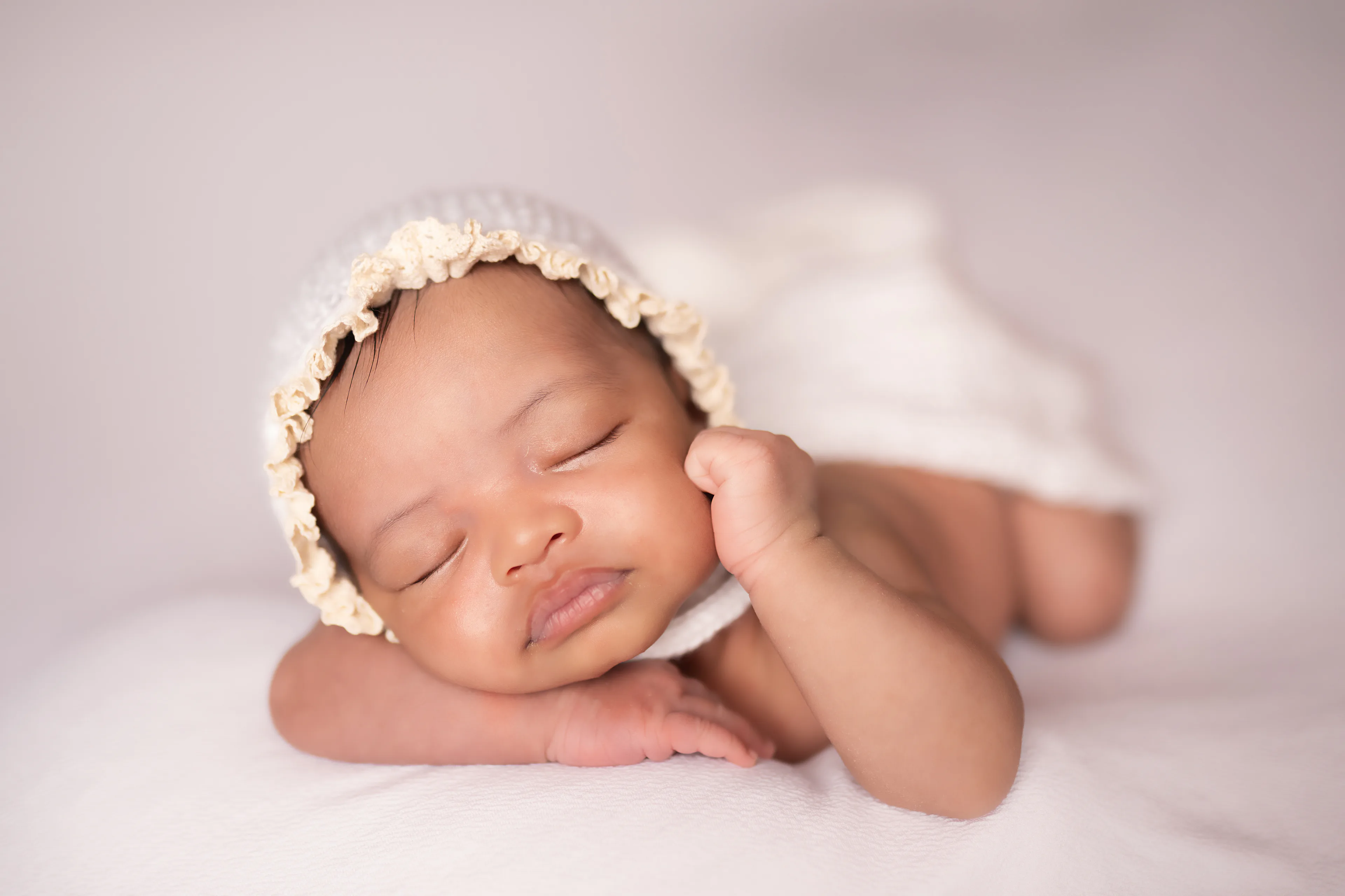 Newborn asleep in a knit bonnet with a white wrap, lying on a cream blanket