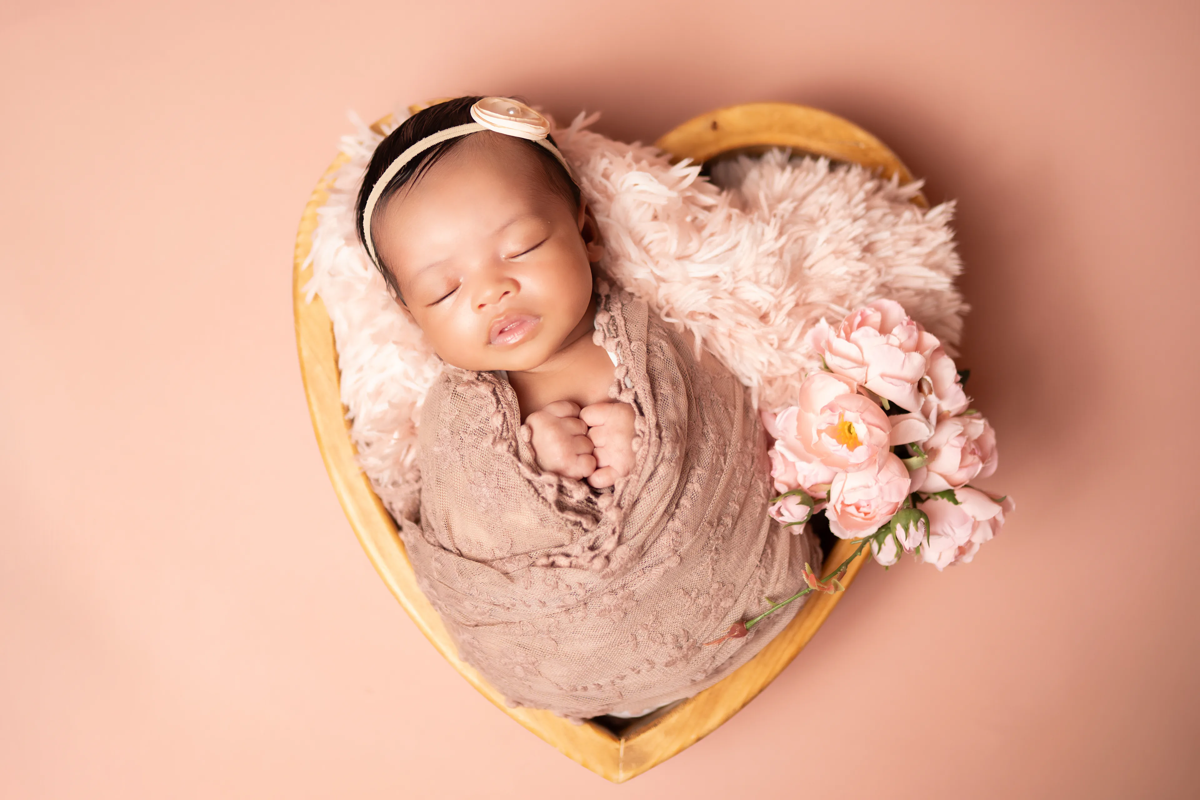 Newborn asleep in a heart-shaped wooden bowl lined with pink fur, on a peach backdrop with blossoms