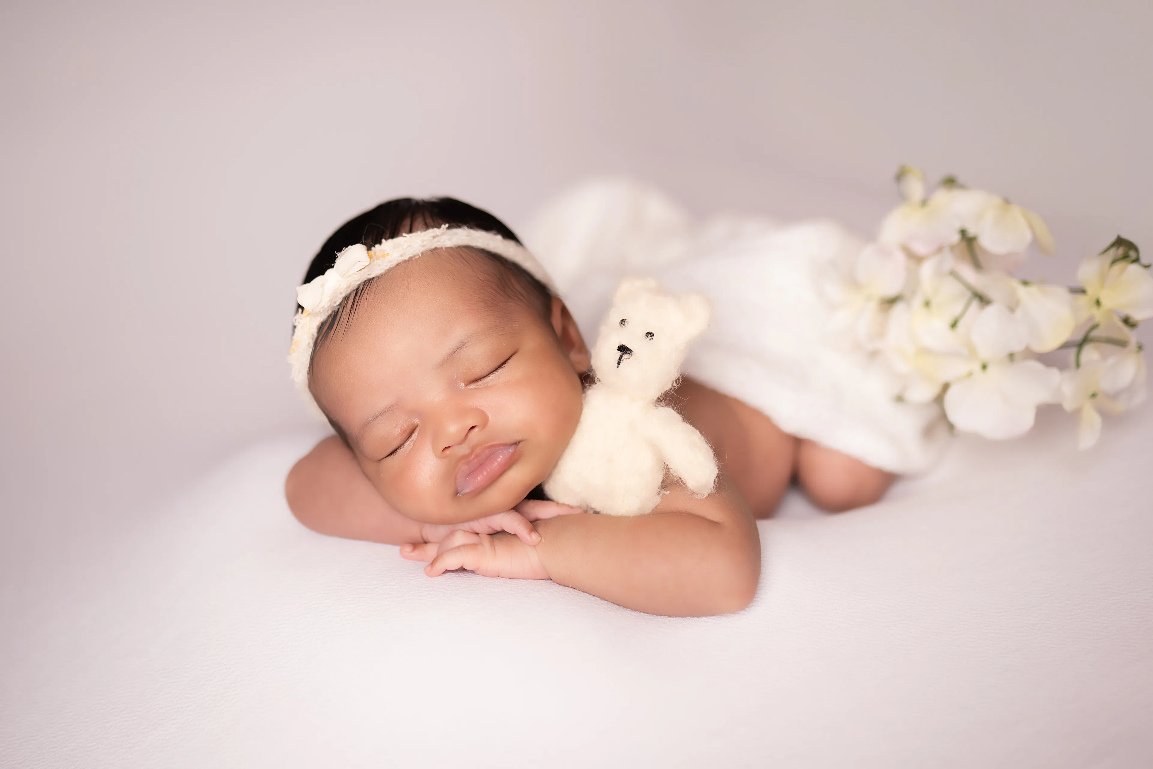 Newborn asleep on a blush backdrop with a small teddy bear and white blossoms beside her