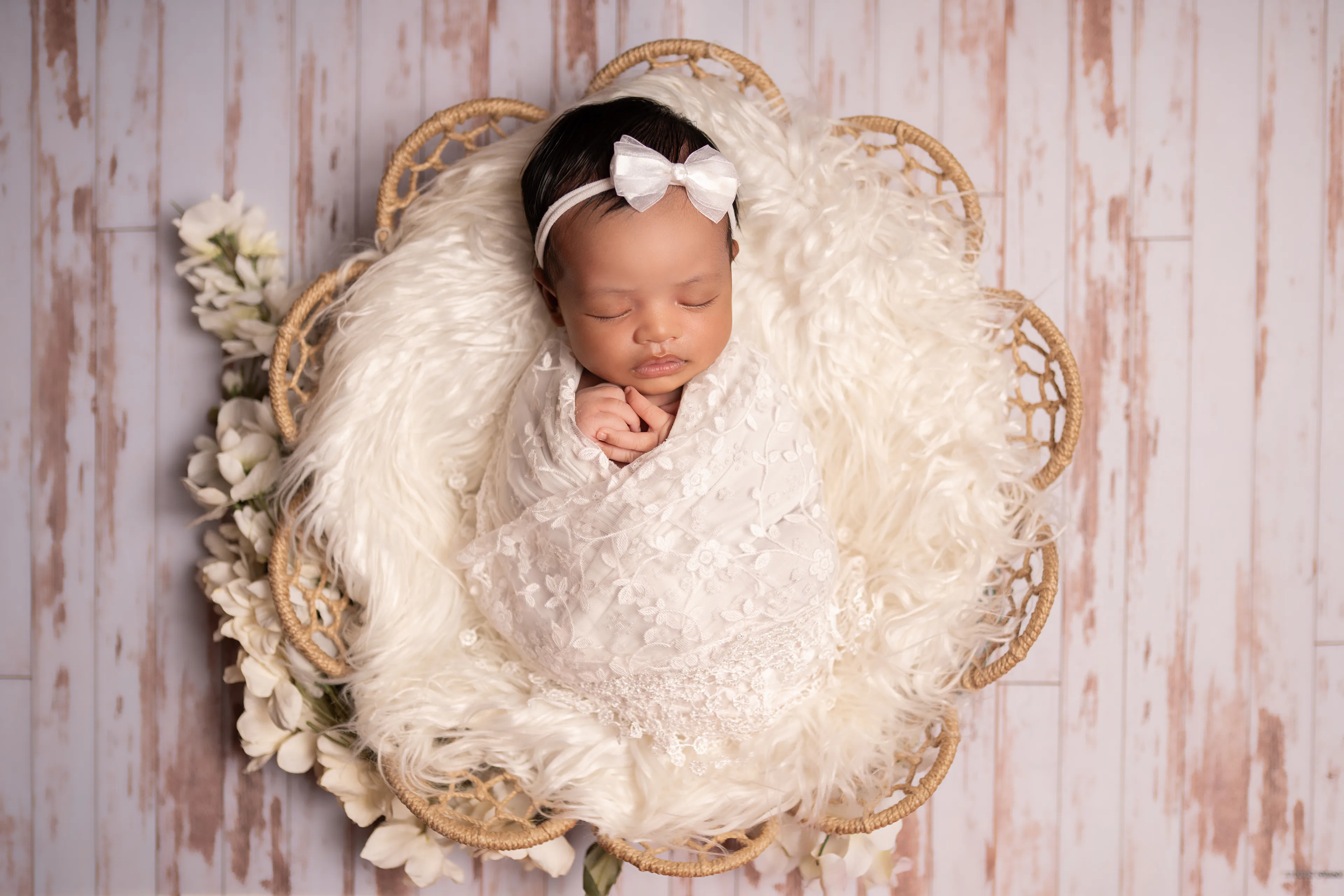 Newborn asleep in a wicker basket lined with cream fur, seen from above, white flowers beside her