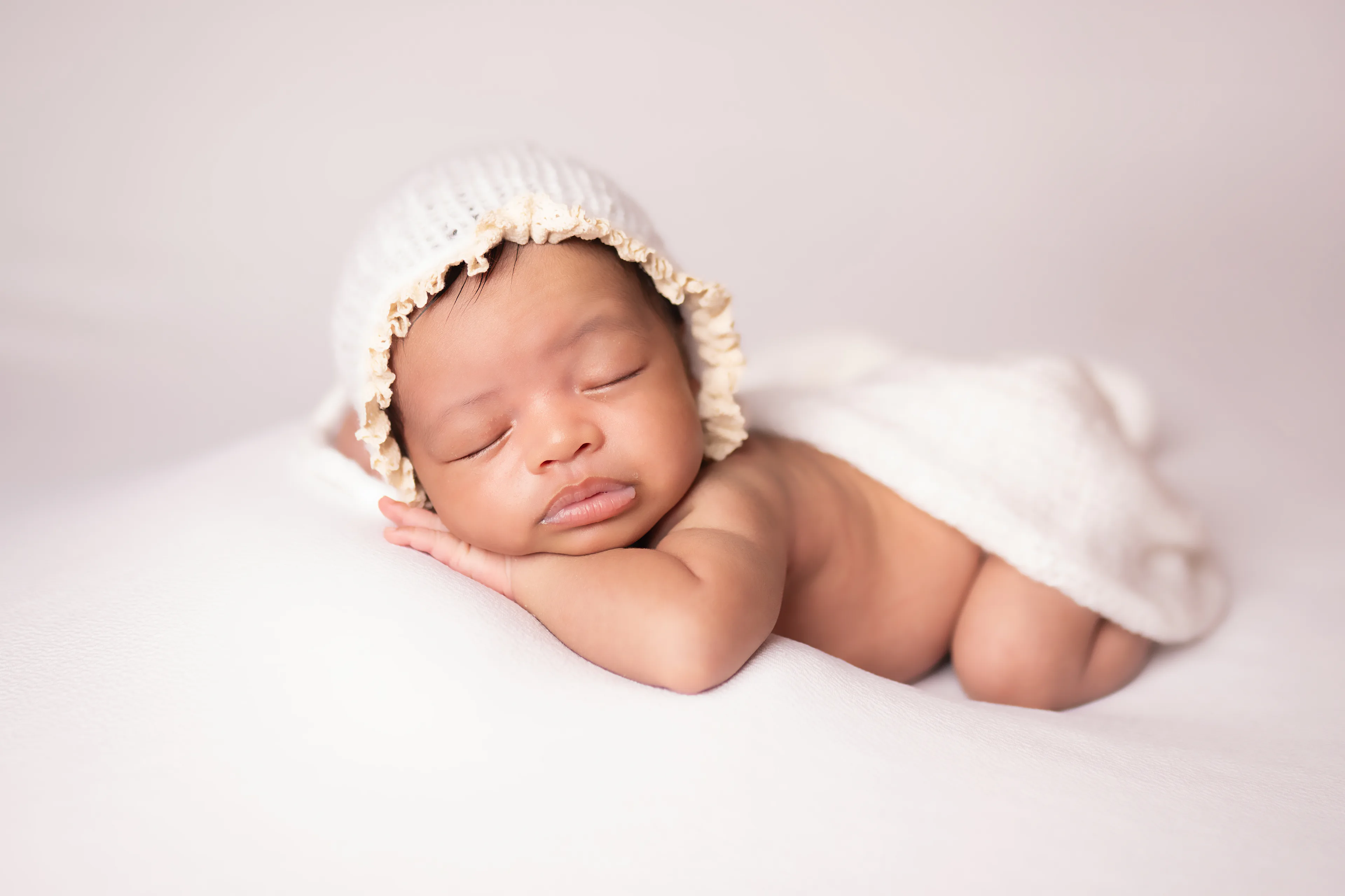Newborn asleep in a floral bonnet, a white wrap over her shoulders, on a soft cream blanket