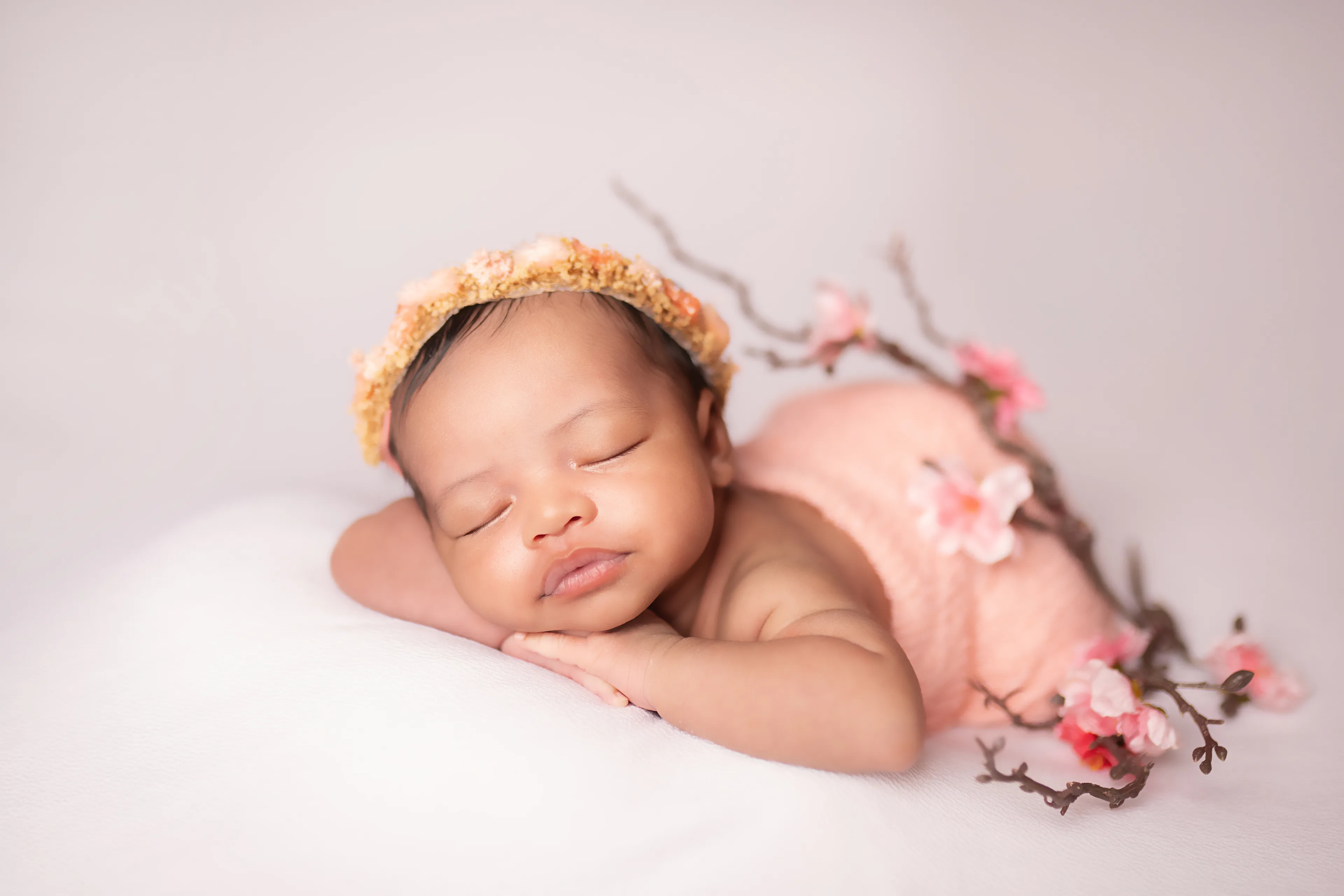 Newborn asleep in a yellow floral crown with a pink wrap and a branch of blossoms beside her