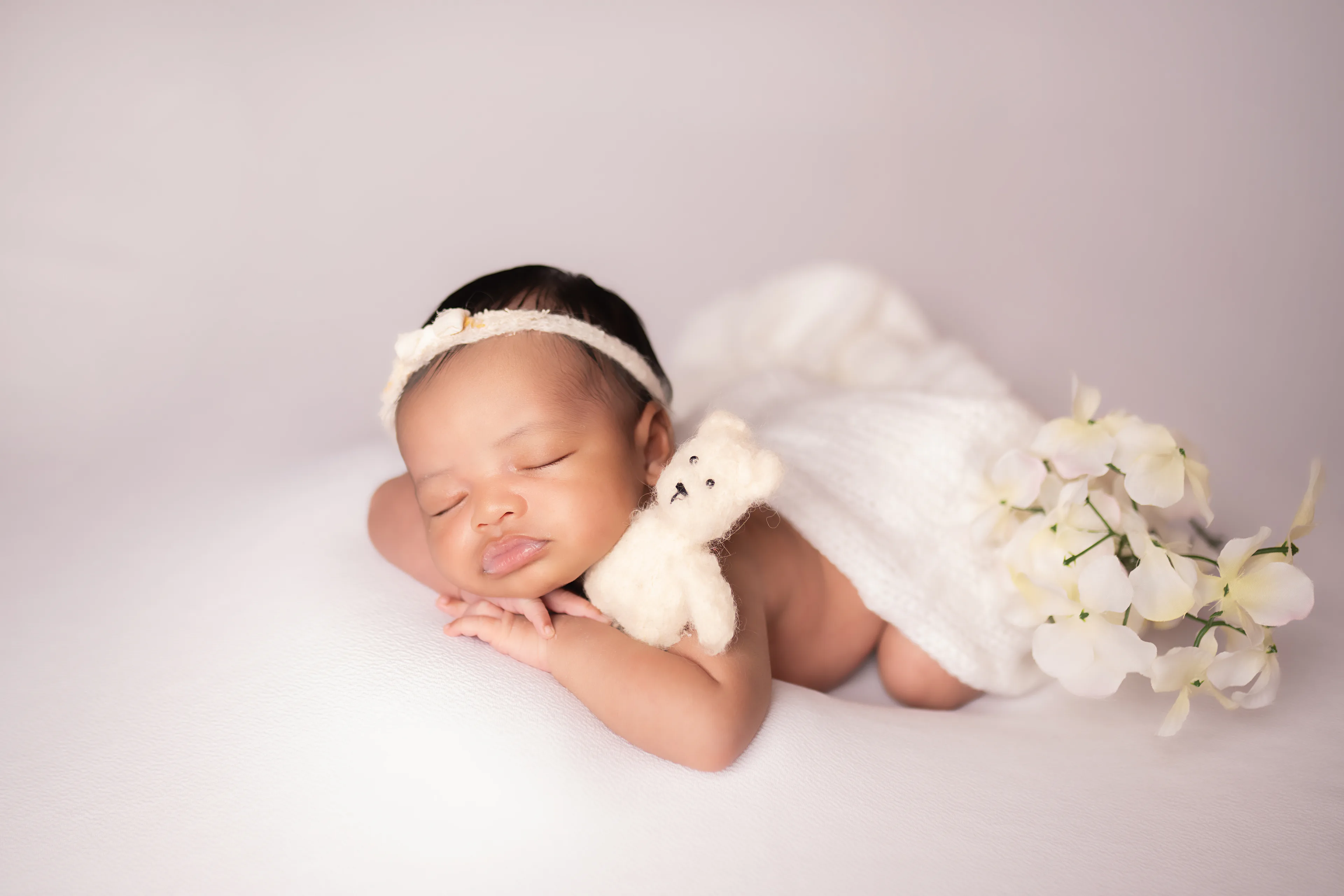 Newborn asleep in a floral bonnet, holding a tiny teddy bear, white blossoms in the background