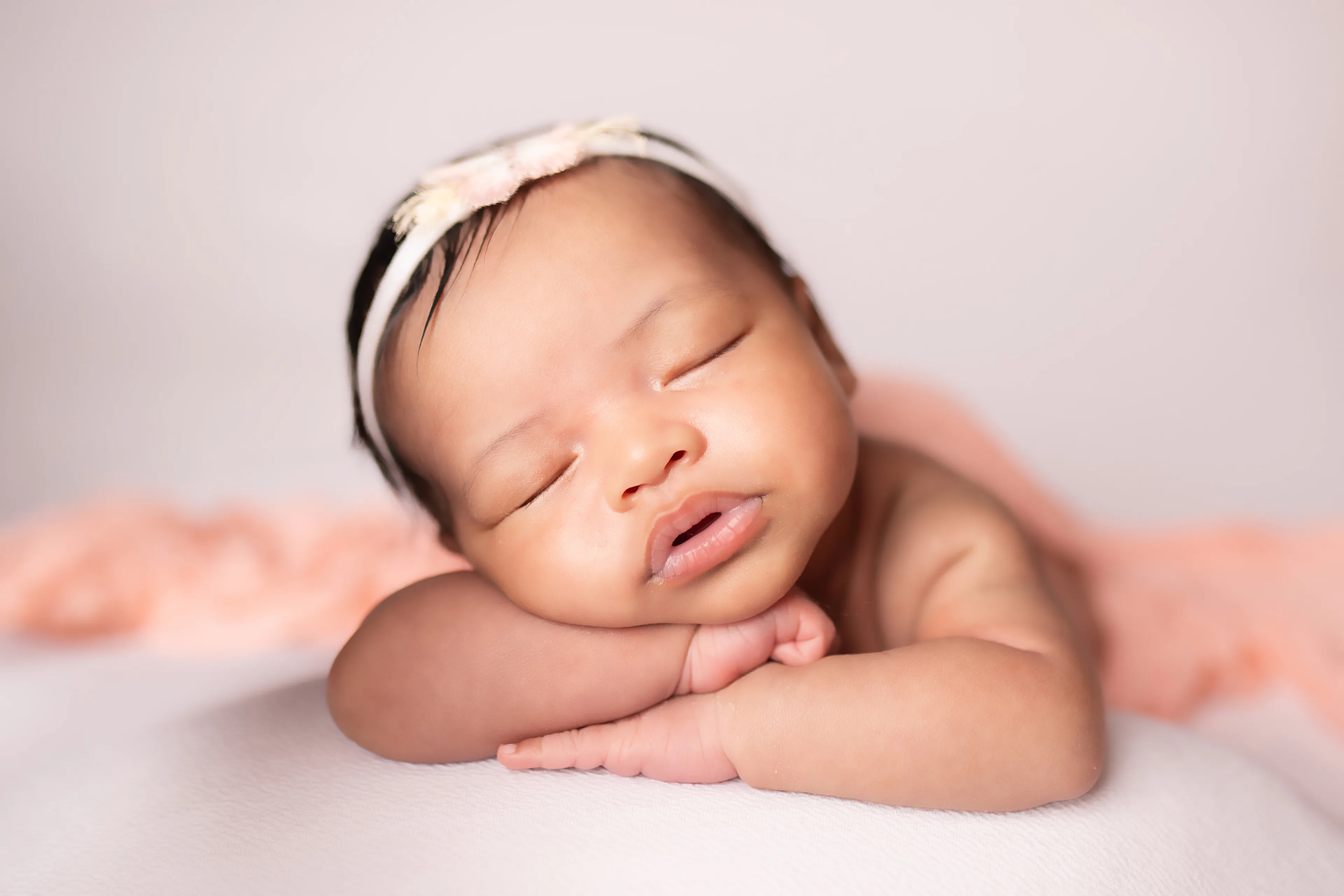 Newborn asleep on a cream blanket with her arms folded under her chin, wrapped in pink