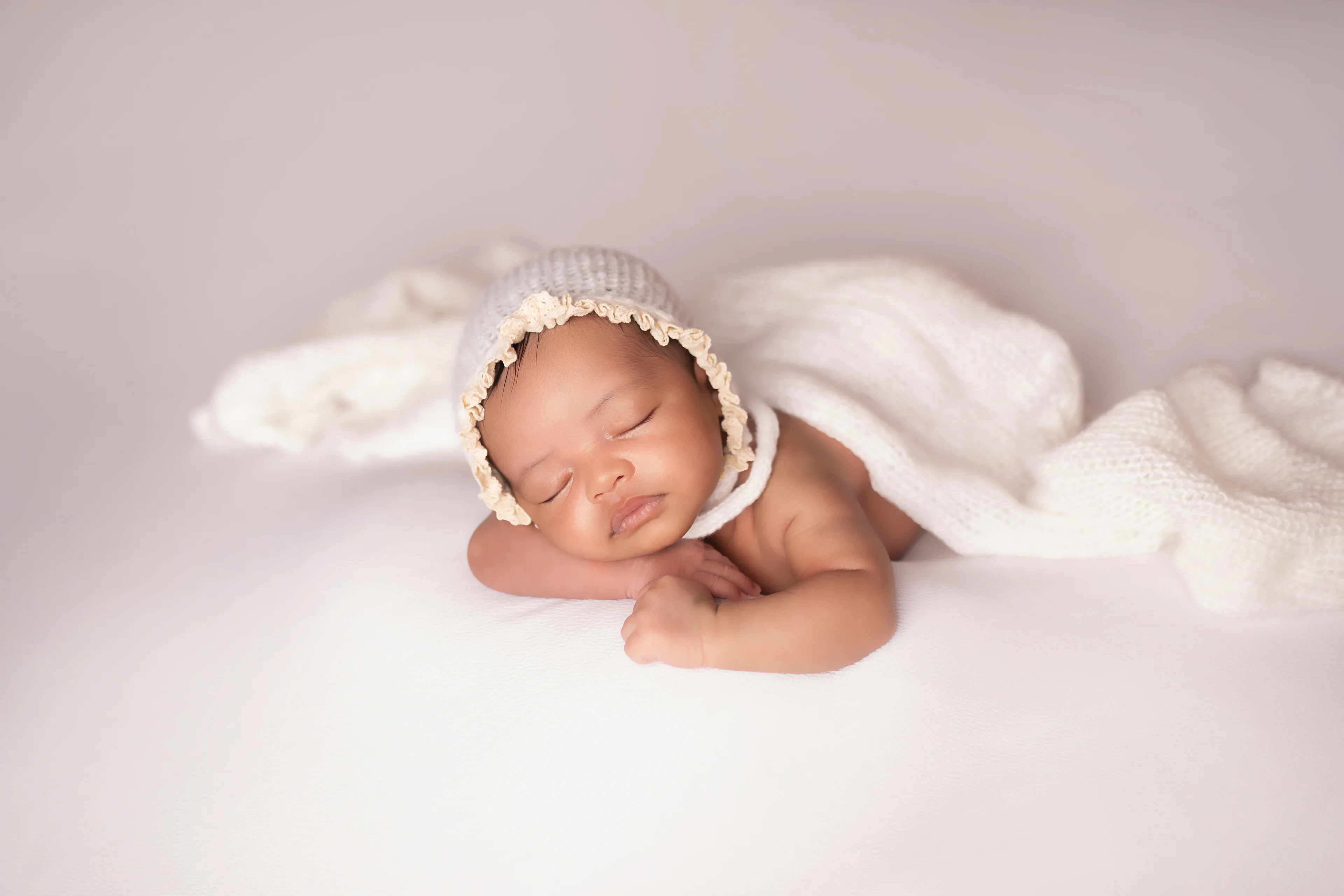 Newborn asleep beneath a white knit wrap on a cream blanket, seen from a distance