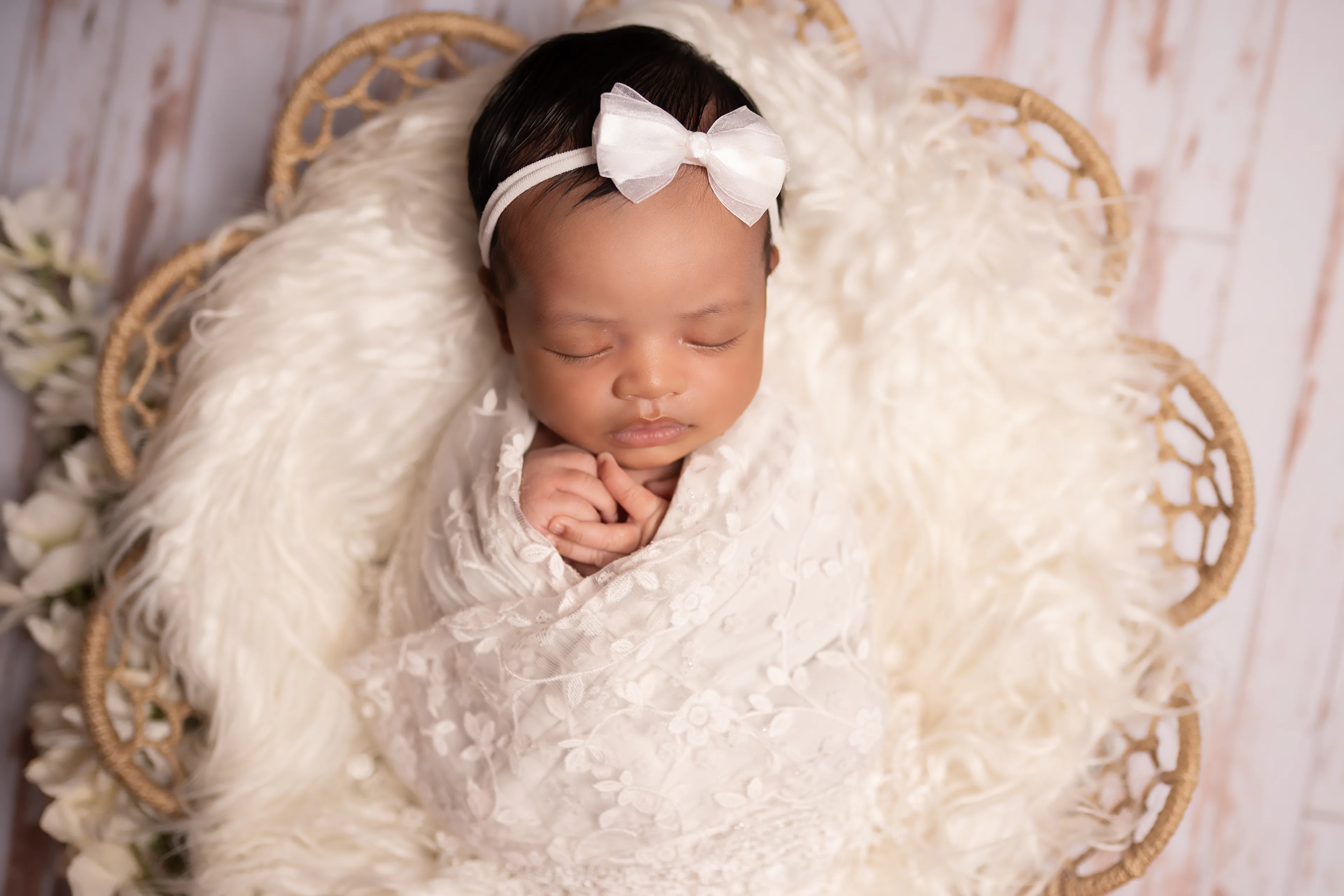 Newborn asleep in a wicker basket with her hands folded, wearing a white bow, on whitewashed wood