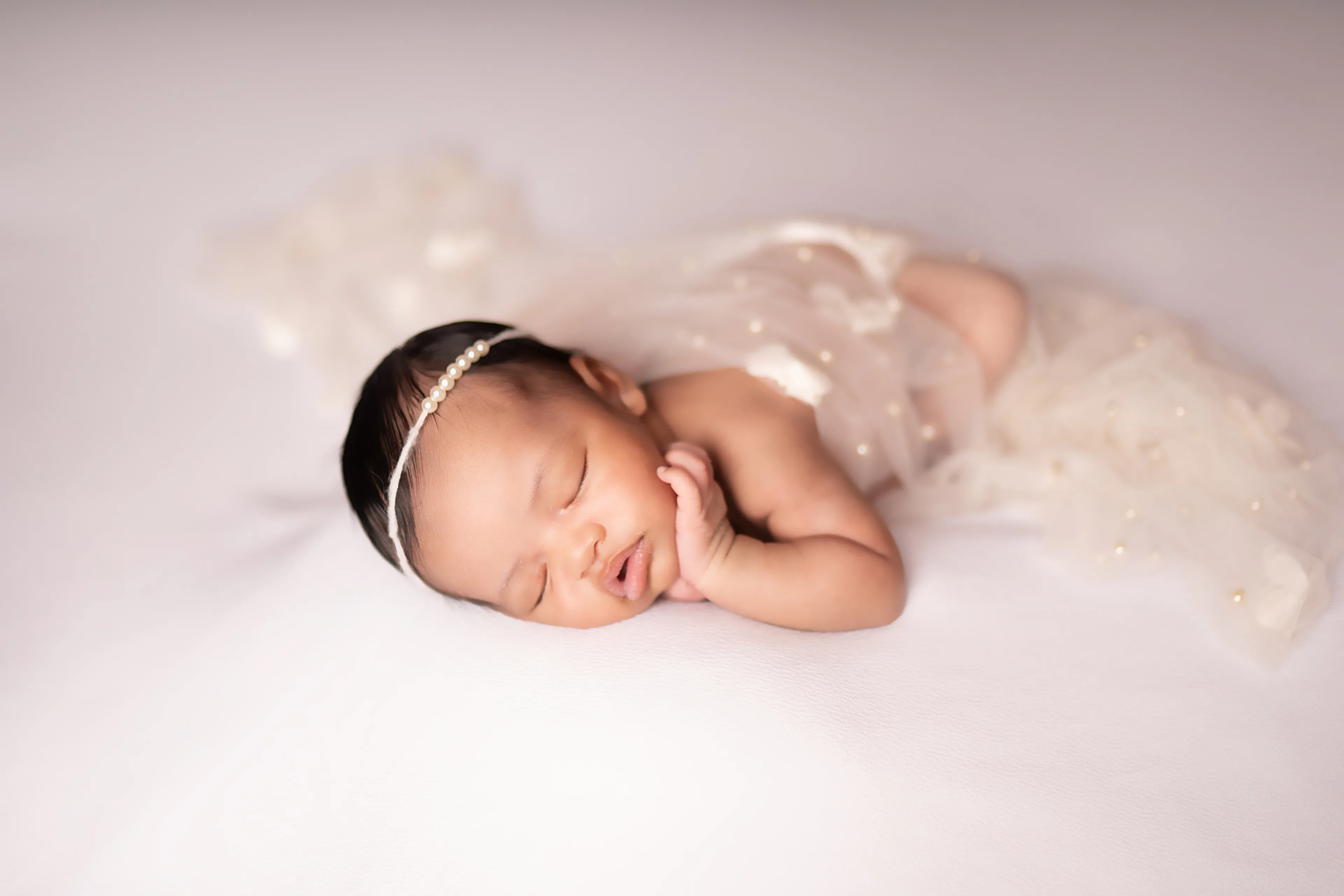 Newborn asleep beneath a sheer pearl-studded veil on a white blanket, seen from a distance